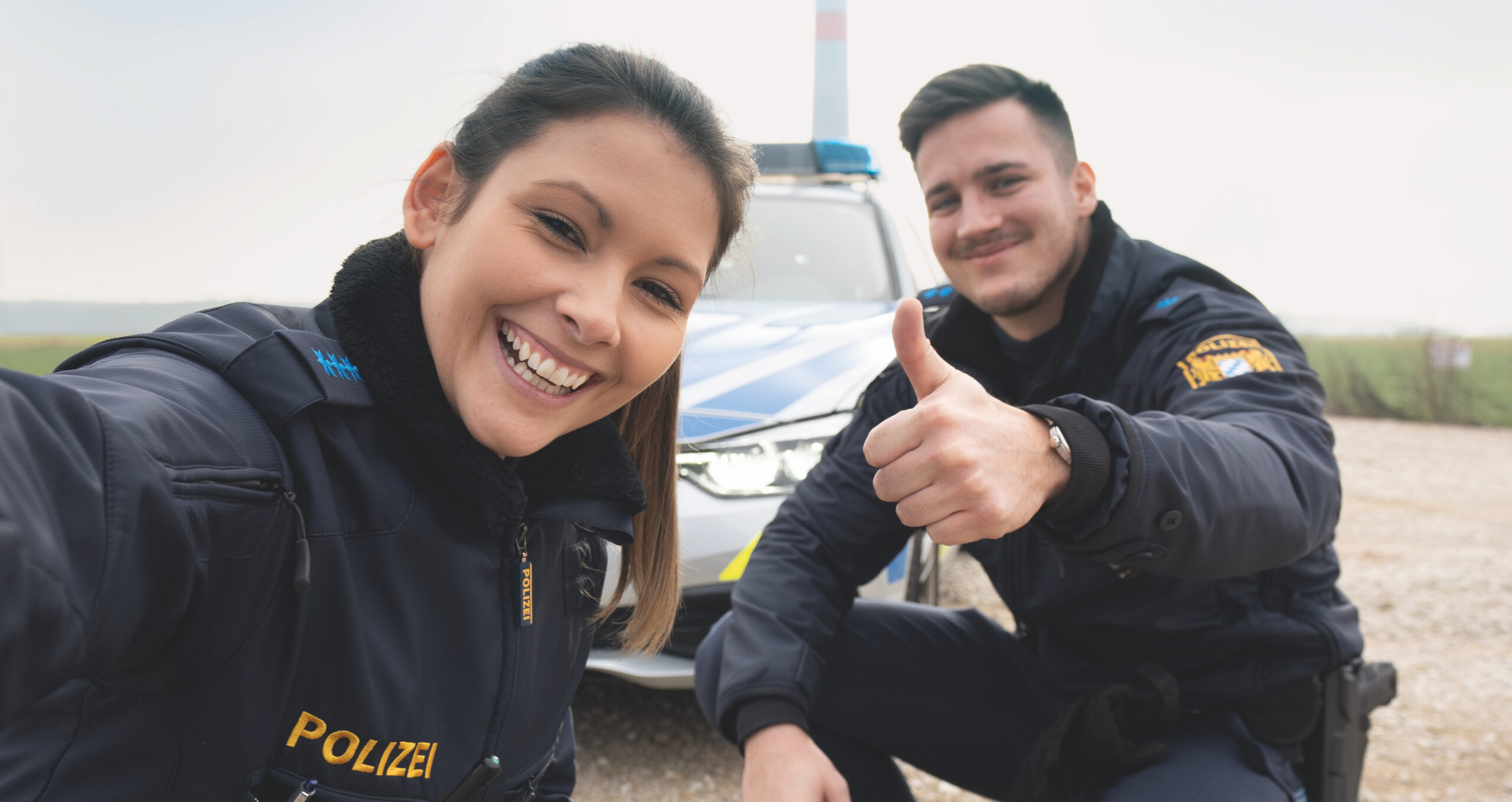 Selfie einer Kollegin und eines Kollegen der Bayerischen Polizei vor ihrem Dienstwagen.